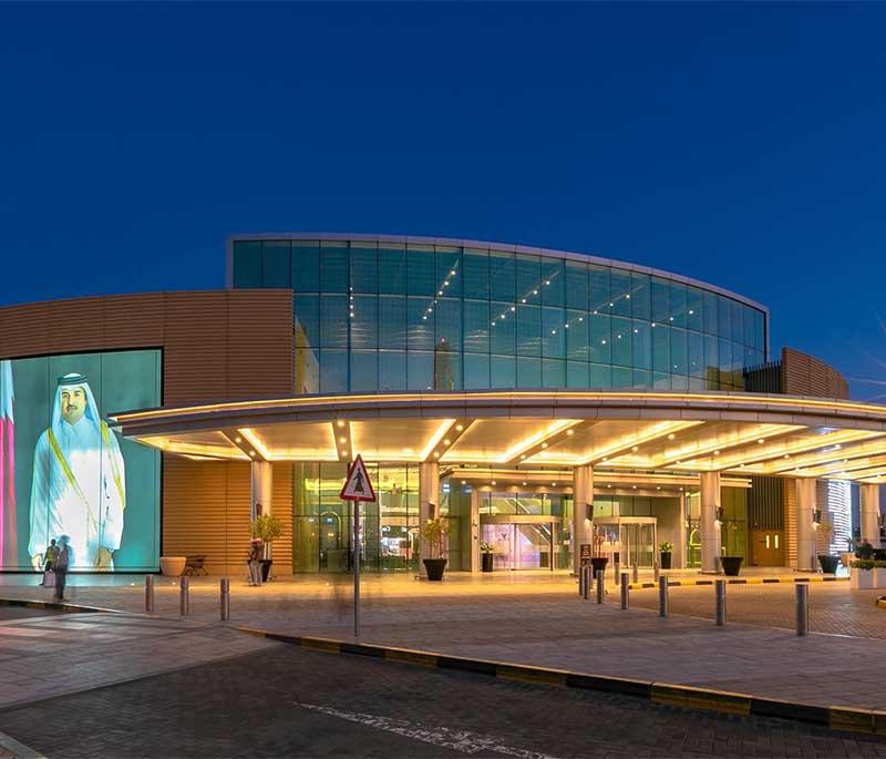 Doha Festival City Mall entrance with bright lights, shoppers entering, and sleek architecture in the evening glow.