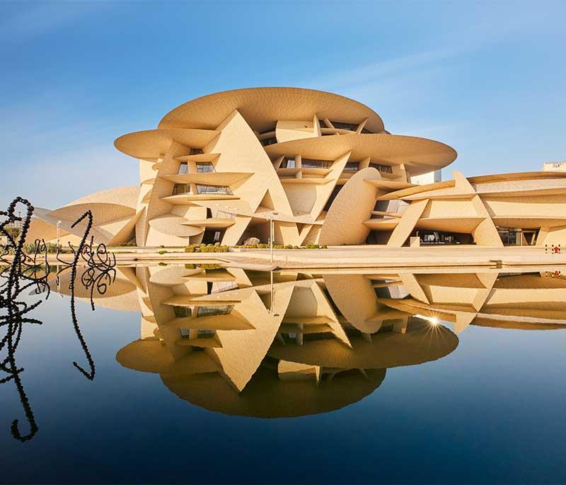 Exterior of the National Museum of Qatar with desert rose–inspired design and tourists exploring the grounds.