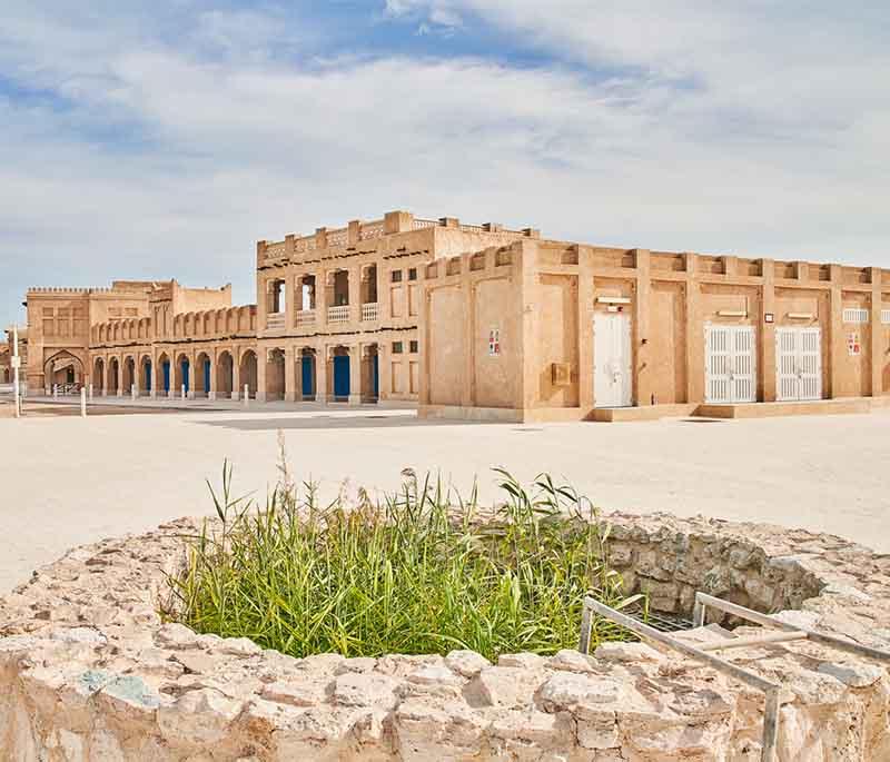 Narrow alleys of Souq Al Wakrah with wooden doors, lanterns, and shoppers exploring the coastal market.